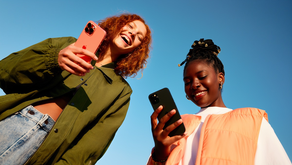 Two friends laughing as they look at their phones outdoors under a blue sky.